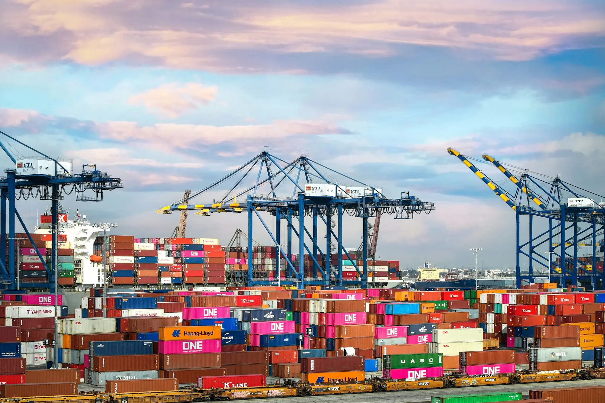 Stacked shipping containers and gantry cranes at a container terminal, waiting on documents before release to the importer